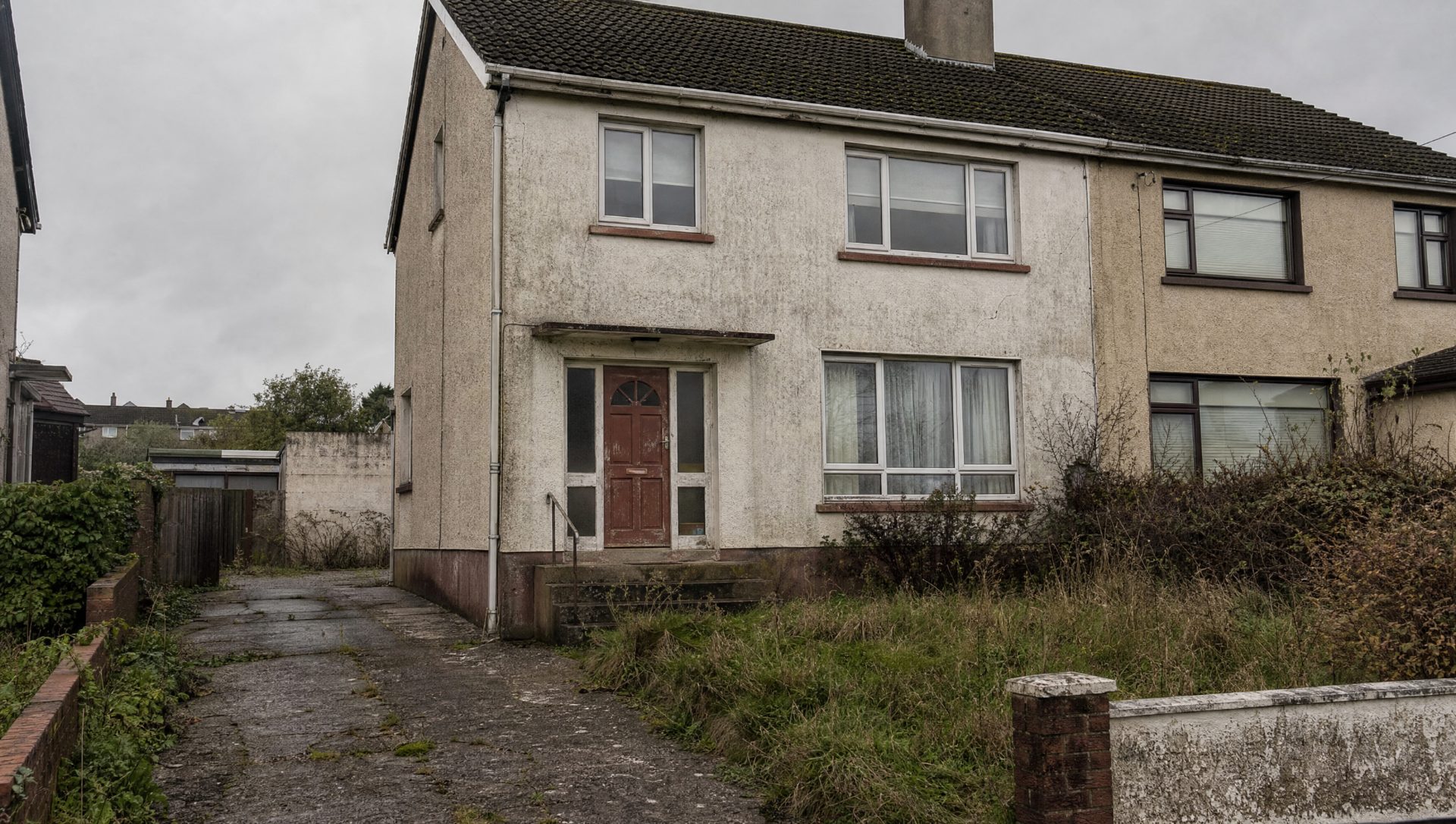 The same house before renovation — dated grey pebble-dash render, worn windows and door, and an overgrown, weedy drive.